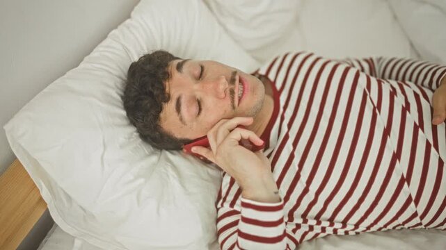 Man lying on bed holds red smartphone to ear, eyes closed and smiling, striped pyjama top, white pillow and wooden headboard in bedroom; relaxed contentment.