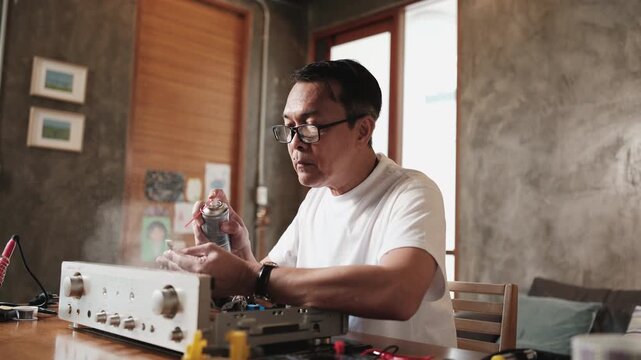 Mature Asian man disassembling and checking audio equipment, spraying contact cleaner, cleaning circuit board for repair, home appliance DIY fix, electronics component hobby, handyman skill workshop.
