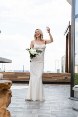 Fototapeta premium Blonde bride joyfully holds a bouquet and a glass, enjoying her wedding day