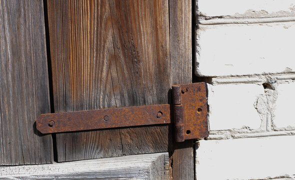 Old rusty metal hinge attached to weathered wooden door and white brick wall close up view. Vintage background with rustic wood texture, corroded iron hinge and old brickwork structure.