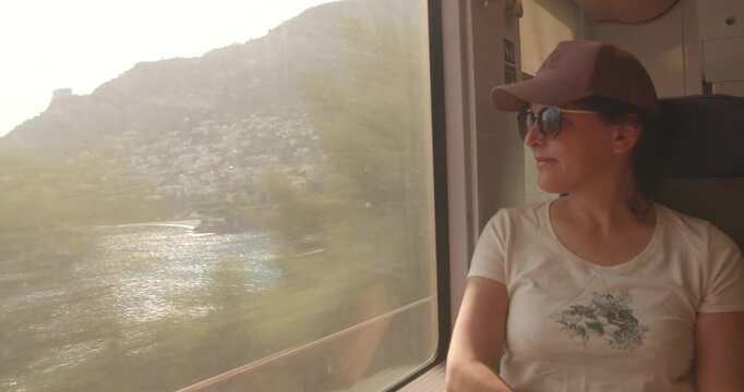 Female passenger wearing cap and sunglasses sits by a train window watching the French Riviera coastline pass in summer light.