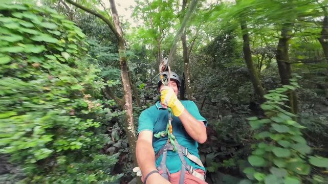 Smiling brown-skinned man films a selfie while zip lining over the lush tropical forest canopy at Constance Ephelia, Mahe Island, Seychelles.