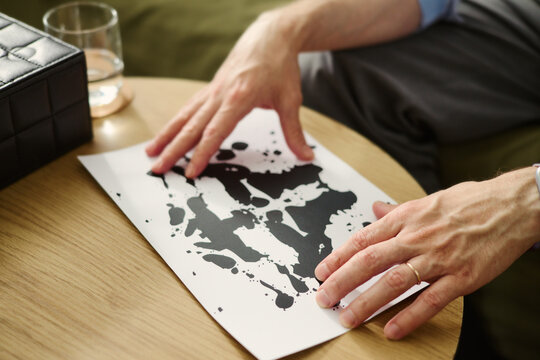 Hands of Caucasian middle aged male psychologist holding Rorschach inkblot test card on wooden table. Image represents mental health assessment, suitable for clinical psychology websites