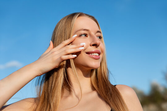 Beautiful woman touching her cheek outdoors with a bright smile and glowing skin, framed against a clear blue sky. Beauty woman portrait presents natural skincare and summer beauty.