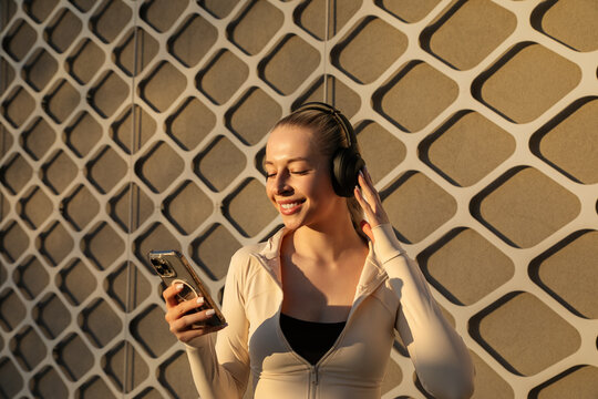 Beautiful woman listening to headphones while checking a smartphone, wearing stylish activewear in warm sunlight. Beauty woman portrait showing modern wellness lifestyle and fitness.