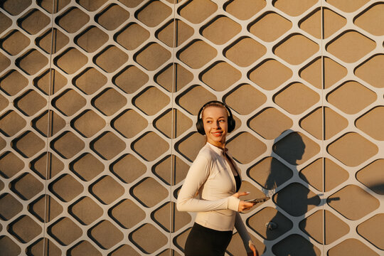 Beauty woman walking with headphones and smartphone beside a geometric wall, wearing sleek activewear in golden light. Attractive woman presents modern fitness and wellness lifestyle.