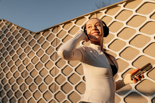 Beauty woman posing with headphones and smartphone against a patterned building, wearing fitted activewear in bright golden light. Beauty woman portrait shows modern wellness lifestyle.