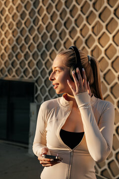 Beauty woman touching headphones while holding a smartphone, wearing fitted activewear in warm outdoor light. Attractive woman portrait presents modern fitness, beauty and wellness lifestyle.