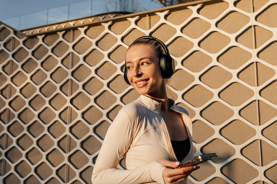 Beautiful woman holding a smartphone while wearing headphones and activewear, smiling beside a geometric building wall in warm light. 