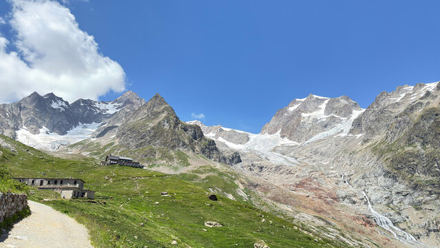 Alpine mountain view rocky peaks glacier green slopes dirt hiking trail stone refuge summer blue sky cloud formations rugged landscape nature scenery outdoors