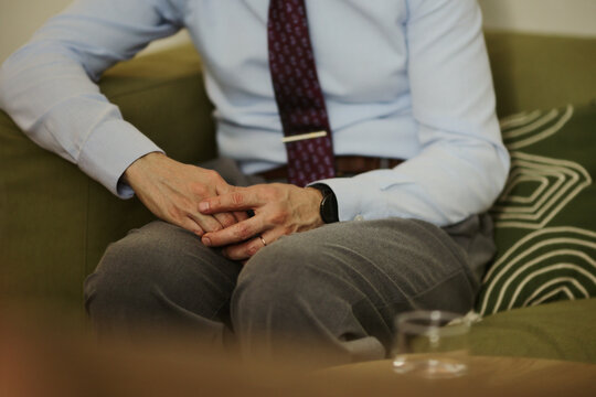 Middle aged Caucasian businessman sitting on sofa with clasped hands during therapy session. Image represents mental health support and is suitable for corporate wellness campaigns