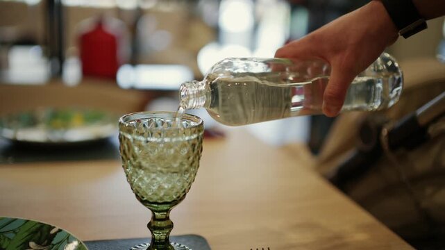 Person pouring clear refreshing water from transparent glass bottle into elegant decorative vintage wine goblet on table.