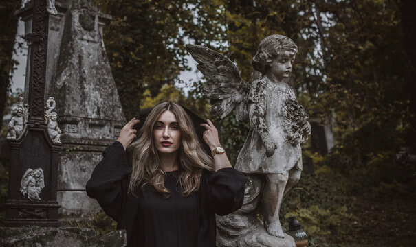 Woman in black hood at old historic cemetery with stone angel statue and ancient crypt, memorial and funerary heritage concept