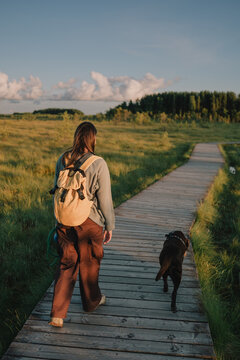 Woman with a backpack walking with her dog along a wooden path through a natural landscape in warm evening light.