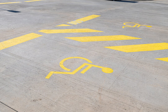 Yellow painted wheelchair accessible parking bay symbol and diagonal hazard stripes on a textured concrete surface under bright natural sunlight in an empty public car park.