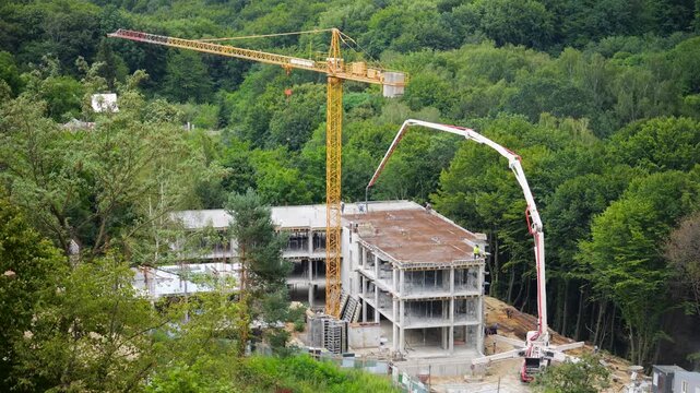 A high-angle view of workers in safety vests operating a yellow tower crane and a concrete pump truck with a long distributor boom, pouring mixture into formwork on a multi-story building near forest.