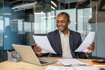Obraz premium Smiling businessman reviewing documents, working at an office desk with a laptop and various paperwork, representing success and diligent professional work