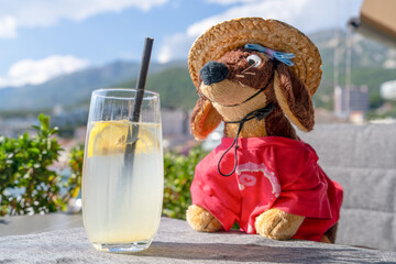 Fototapeta premium A stuffed dachshund dog toy wearing a straw hat and red shirt sits beside a tall glass of lemonade with lemon slices on an outdoor table overlooking mountains, a concept of summer vacation relaxation.