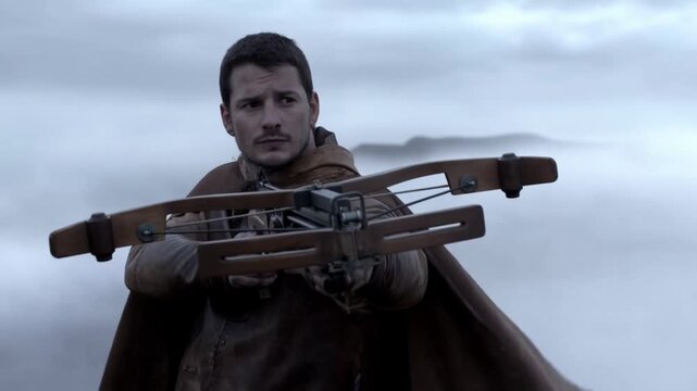 Tracking shot man aiming large crossbow while turning on foggy moorland with flowing cloak under overcast sky tense stance preparing to fire