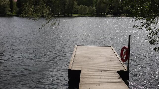 A weathered wooden dock extends over a sparkling lake in Lahti, framed by lush green trees and a bright blue sky dotted with fluffy clouds on a sunny summer day.