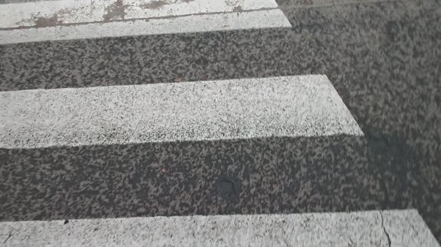 POV looking down as a pedestrian steps off the curb and walks across a faded zebra crosswalk on a city street.