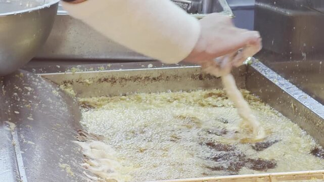 Commercial kitchen view of a chef preparing traditional tempura. Battered strips of seafood or vegetables are carefully dipped into a deep stainless steel fryer filled with hot, bubbling oil and