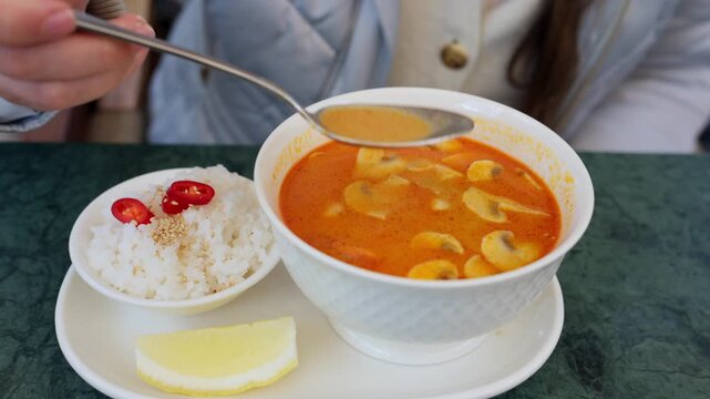 Woman eats tom yum soup with shrimp and mushrooms from white bowl.