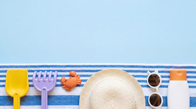Flat lay composition of children's beach toys, straw hat, sunglasses, and sunscreen lotion on a striped towel against a blue background, with copy space.