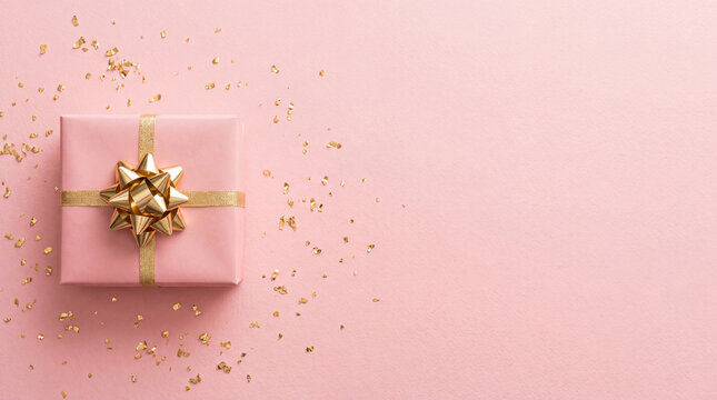 A festive pink gift box tied with a shiny gold ribbon, surrounded by golden confetti flakes on a matching pink background with copy space.