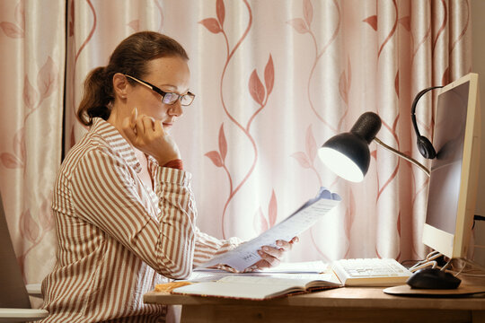 Focused middle-aged woman in glasses sitting home desk and reading paper documents under desk lamp near computer monitor in evening.