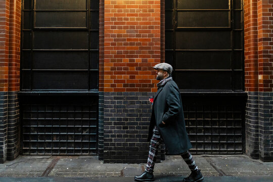 Male in winter coat walks past brick building holding coffee cup