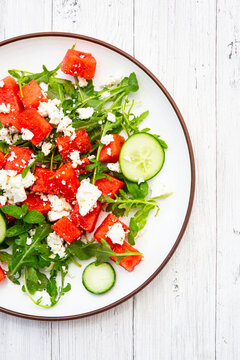A fresh watermelon salad with feta and arugula sits on a wooden table