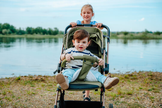 A smiling girl pushes her baby brother in a stroller by the pond