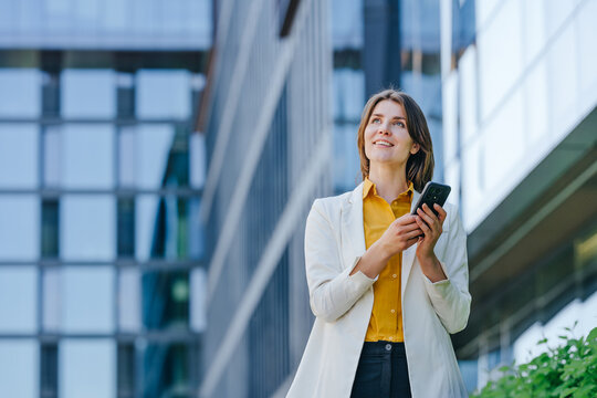 Smiling businesswoman in formal wear holds a phone outdoors
