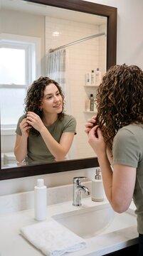 Young curly haired woman styling her hair in front of bathroom mirror with hair care morning routine self care personal hygiene bathroom
