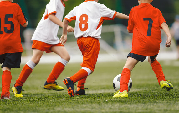 Junior Soccer Players in Orange Kits Dribbling Ball During Competitive Match - Youth Football Players Number 7, 8, and 15 Running on Pitch