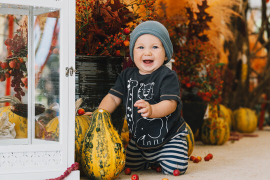 A happy baby boy wearing a grey beanie sits outdoors next to a white vintage lantern and a large orange pumpkin. Joyful childhood moments celebrating the beautiful autumn season.