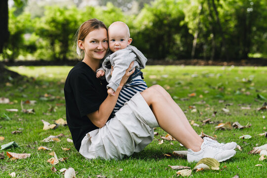 A young happy mother sits on the green park grass holding her lovely baby boy on a warm autumn day. Authentic family lifestyle representing tender parenting and outdoor recreation.