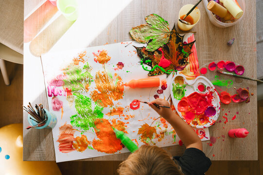Top down view of messy children's art activities on a wooden table with bright paints and autumn leaves. Creative early education and fun messy art projects for preschool toddlers.