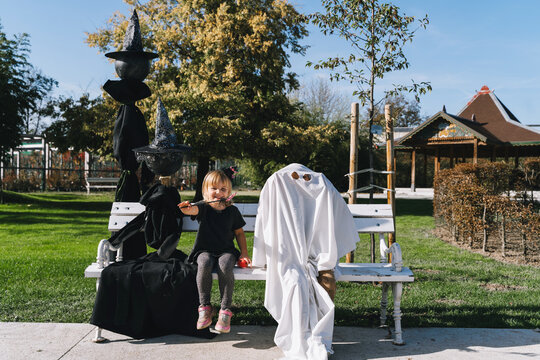 A young girl sits on a white park bench next to a spooky white ghost decoration and black witch hats. Joyful outdoor Halloween preparation and fun seasonal autumn lifestyle concept.