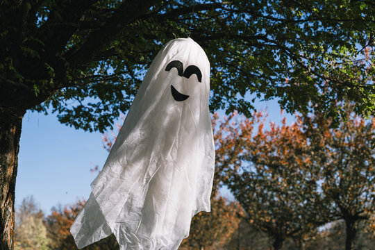 A large handmade white ghost decoration with a funny face hangs from a beautiful green tree in a public park. Spooky seasonal Halloween preparation and festive outdoor lifestyle.