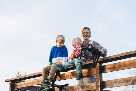 A smiling mother and two young sons sit on a rustic wooden fence playing with a large black toy spider. Joyful family Halloween preparation and fun seasonal outdoor lifestyle.