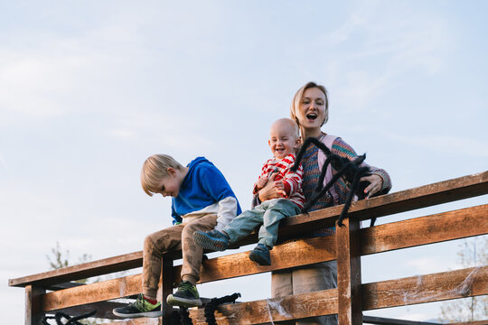 A smiling mother and two young sons sit on a rustic wooden fence playing with a large black toy spider. Joyful family Halloween preparation and fun seasonal outdoor lifestyle.