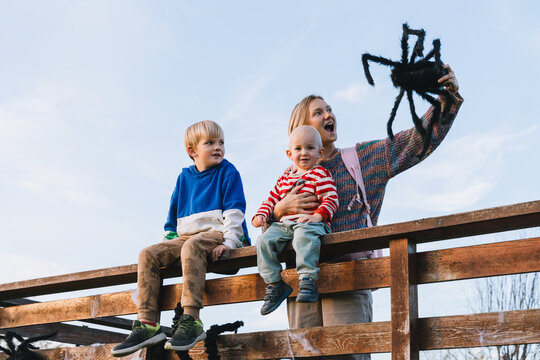 A cheerful mother lifts a giant black toy spider high in the air while sitting on a wooden fence with her two little sons. Playful family outdoor lifestyle during Halloween time.