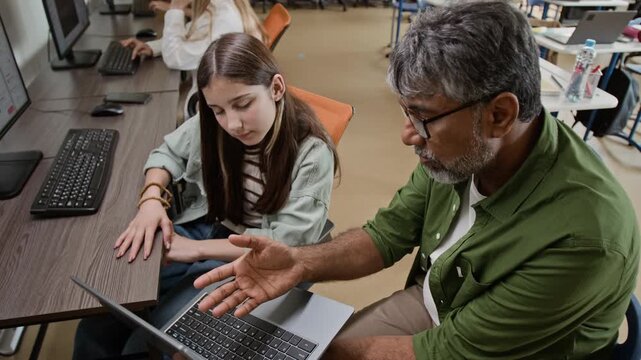 Middle aged South Asian teacher explaining software on laptop to Caucasian teenager girl in computer class, high angle shot. Footage represents STEM education marketing and e-learning promos