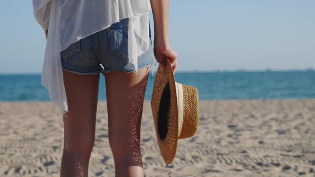 Rear view of young woman, standing, hold hat, beach on sunny summer day, wearing casual denim shorts, enjoying tropical holiday, selective focus on straw accessory and ocean.