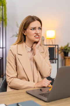 Middle-aged businesswoman at home office works on laptop feels tooth pain winces holds cheek tries to stay focused. Freelancer girl at table keeps typing slow pace rubs jaw pauses continues report on