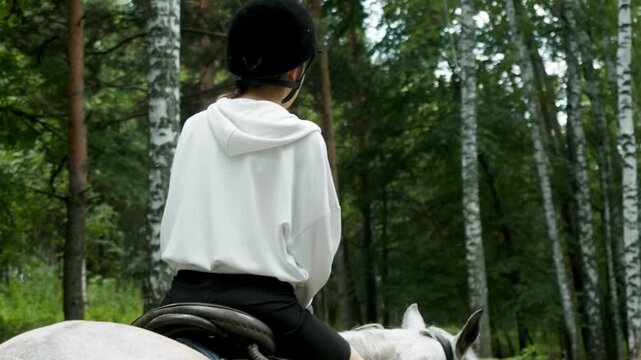 Female instructor leads gray horse with young rider through birch forest. Rear view shows supervised horseback riding lesson on woodland trail.