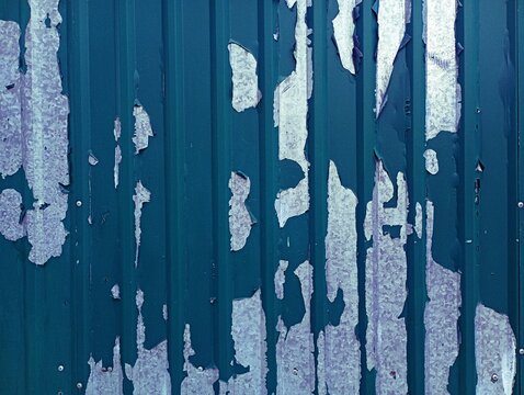 Texture of a damaged corrugated metal sheet wall with peeling teal paint revealing weathered galvanized steel underneath.