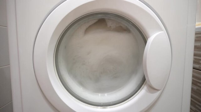 Modern washing machine with laundry inside, view through the glass door, household appliances in the process of cleaning